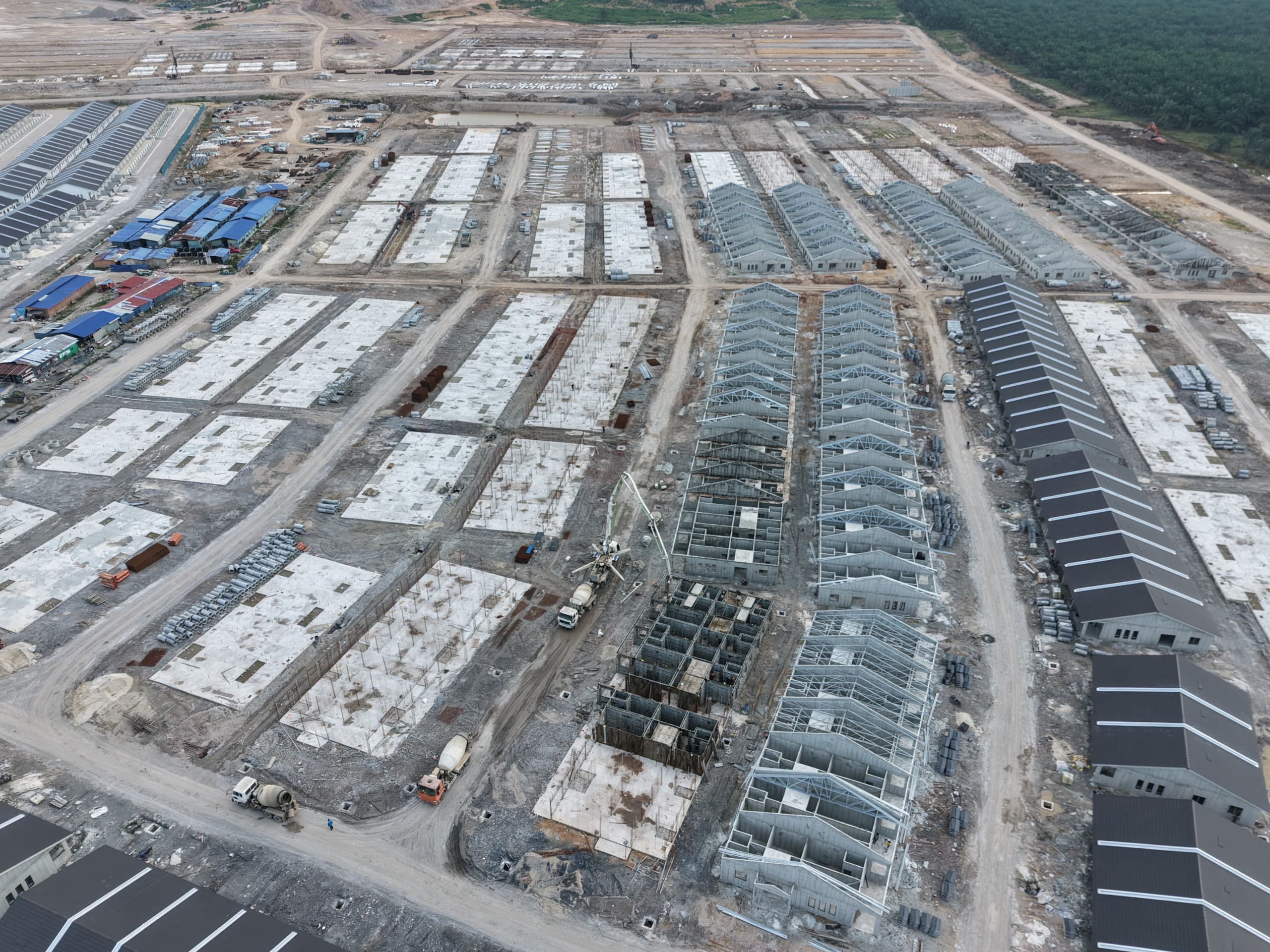 Aerial view of concrete slabs, exposed roof frames, dark-roofed rows and construction vehicles.