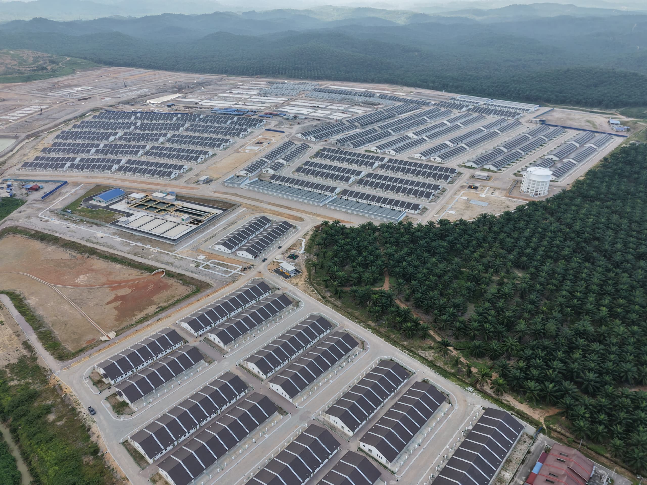Aerial view of dark-roofed housing rows, paved roads, white cylindrical tower and forested hills.